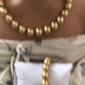 May include: A gold-toned beaded necklace featuring a series of uniform, smooth, spherical beads arranged in a single strand, paired with a matching gold-toned beaded bracelet displayed on a small, rectangular white fabric cushion.