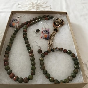 May include: A jewelry set featuring a necklace and bracelet made of round green and red unakite beads, a gold-tone lion head pendant with a chain tassel, and two pairs of dangling earrings with cat illustrations wearing hoodies and drinking boba.
