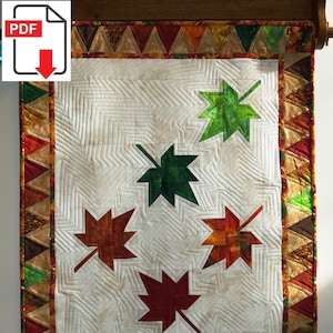 Könnte beinhalten: Eine weiße Steppdecke mit einem Patchwork-Rand aus braunen, roten und grünen Dreiecken. Die Steppdecke zeigt sechs aufgenähte Ahornblätter in verschiedenen Grüntönen, Braun- und Rottönen.