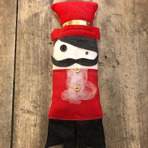 Può includere: Un pupazzo di feltro con un cappello rosso, un baffo nero e un fiocco bianco. Il pupazzo indossa una giacca rossa con un bottone dorato e pantaloni neri.