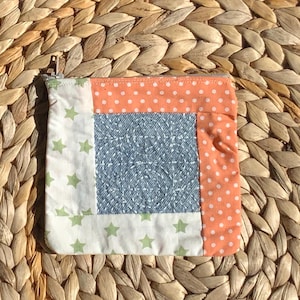 May include: A small, square patchwork pouch with a zipper closure. The pouch features a central denim-like square, bordered by white fabric with green stars, and orange fabric with white polka dots. The pouch is set against a woven background.