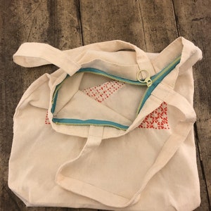 May include: A white canvas tote bag with a zipper closure and red embroidered details. The zipper is teal blue and has a gold pull.