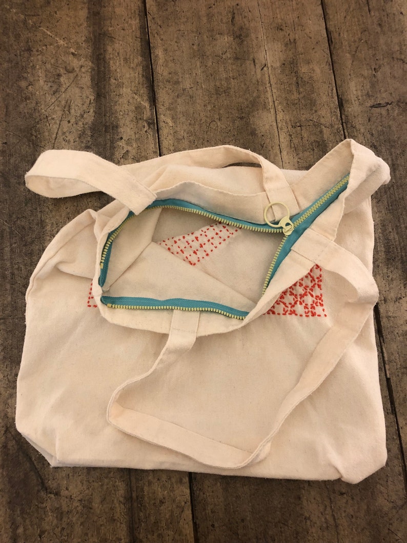 May include: A white canvas tote bag with a zipper closure and red embroidered details. The zipper is teal blue and has a gold pull.