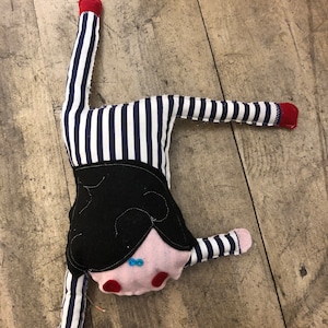 May include: A handmade doll in a handstand pose. The doll has a pink face with red cheeks and blue button eyes. It wears a black felt top and striped blue and white fabric legs and arms. The feet and hands are red.