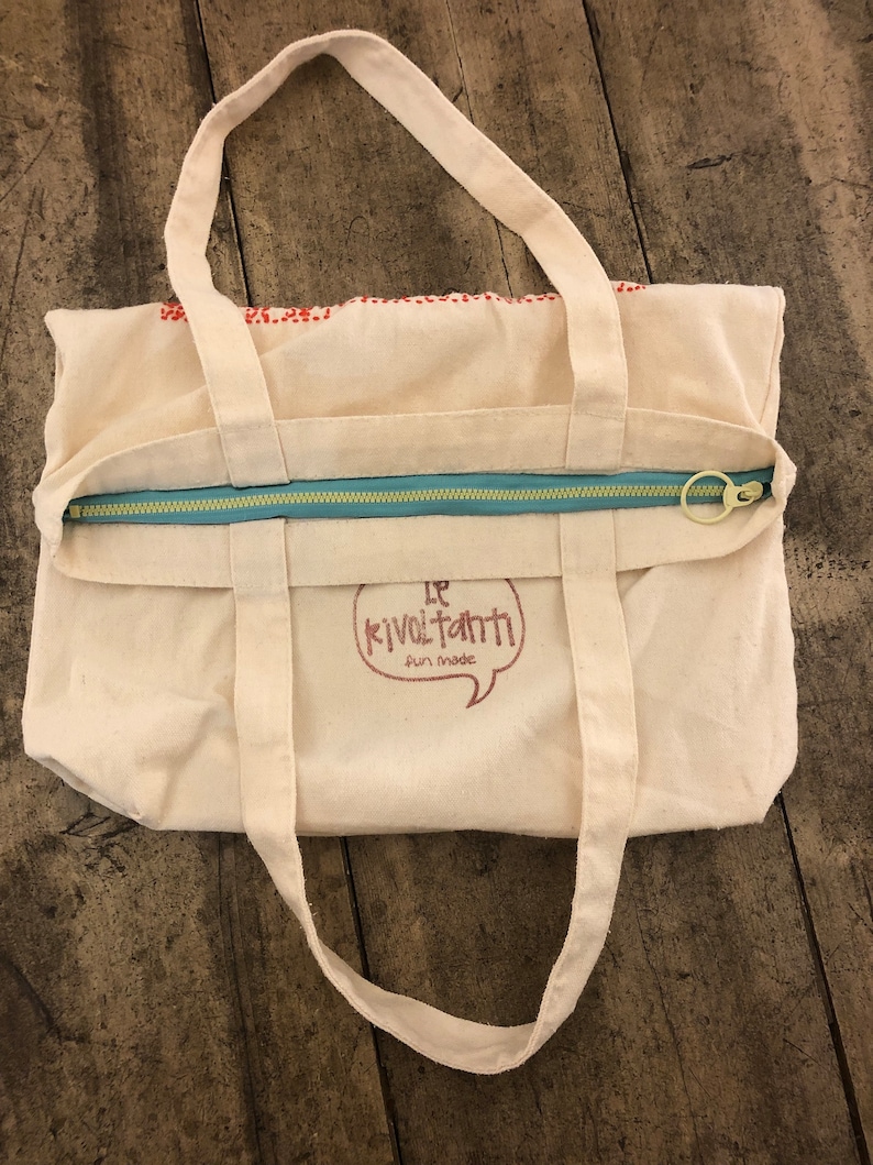May include: A white canvas tote bag with a zipper closure and two handles. The bag has a red and white logo that says "Le Kivoltanti Fun Made".