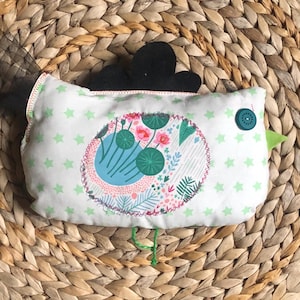 May include: A white bird-shaped pillow with green star patterns and a floral design. The pillow has a black tulle tail, a green button eye, and a green beak. The pillow is on a woven wicker surface.