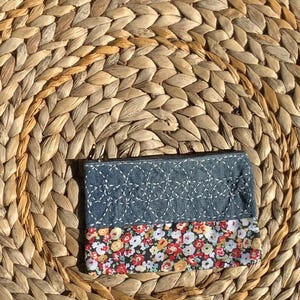 May include: A small, rectangular denim and floral print pouch with a zipper closure. The top half is a blue denim with white stitching, and the bottom half features a floral pattern with red, yellow, and white flowers on a black background.