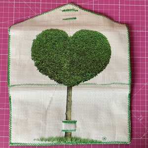 May include: A white fabric envelope with a green heart-shaped tree design. The envelope has a green stitched border and a small white label with a registered trademark symbol.