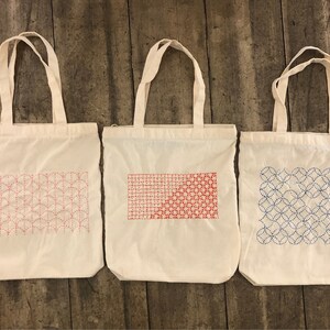 May include: Three white canvas tote bags with different embroidered designs. The first bag has a red and pink geometric pattern, the second bag has a red and pink geometric pattern, and the third bag has a blue geometric pattern.