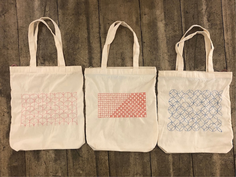 May include: Three white canvas tote bags with different embroidered designs. The first bag has a red and pink geometric pattern, the second bag has a red and pink geometric pattern, and the third bag has a blue geometric pattern.