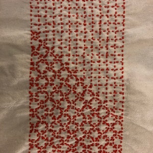 May include: A white fabric with a red embroidered design. The design is a grid pattern with a repeating floral motif.