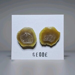May include: Two geode slices with a yellow and white outer edge and a sparkling crystal center. The word "GEODE" is printed in black block letters below the geodes on a white card.