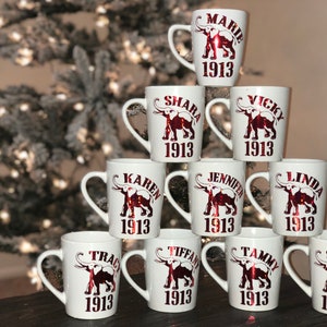 May include: White ceramic mugs with names and the year 1913 printed in red. Each mug features an elephant graphic. The mugs are stacked in a pyramid shape, with a blurred Christmas tree in the background.