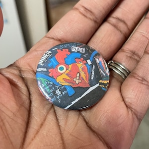 May include: A round button with a black background and a colorful graphic of a heart with a chicken head and a bandage on it. The text "INNER" and "PEACE" are visible on the button.