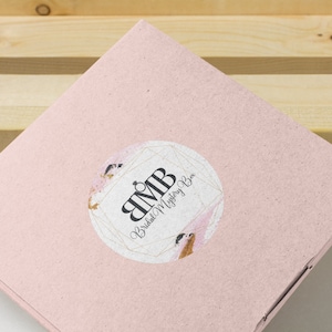 Puede incluir: Una caja de cartón rosa con una pegatina redonda que presenta un diseño geométrico dorado y el texto "BMB Bridal Mystery Box".