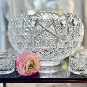 May include: A clear, cut-glass punch bowl with matching small cups. The bowl has a scalloped rim and sits on a pedestal base. A pink flower rests near the bowl and cups. The glassware is likely vintage.