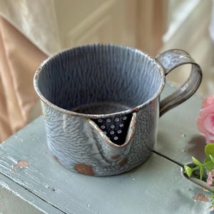 May include: A vintage, blue and white enamelware pitcher with a handle and spout. The pitcher has a textured pattern and shows signs of wear, with rust-colored spots. A pink rose and greenery are in the background.