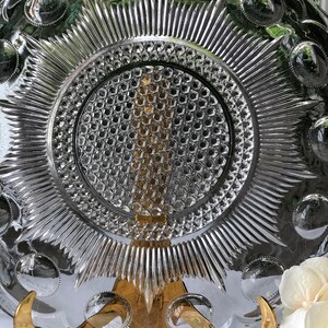 May include: A clear glass plate with a raised, starburst design and a central, circular, honeycomb pattern. The plate has a scalloped edge with raised circles around the rim.
