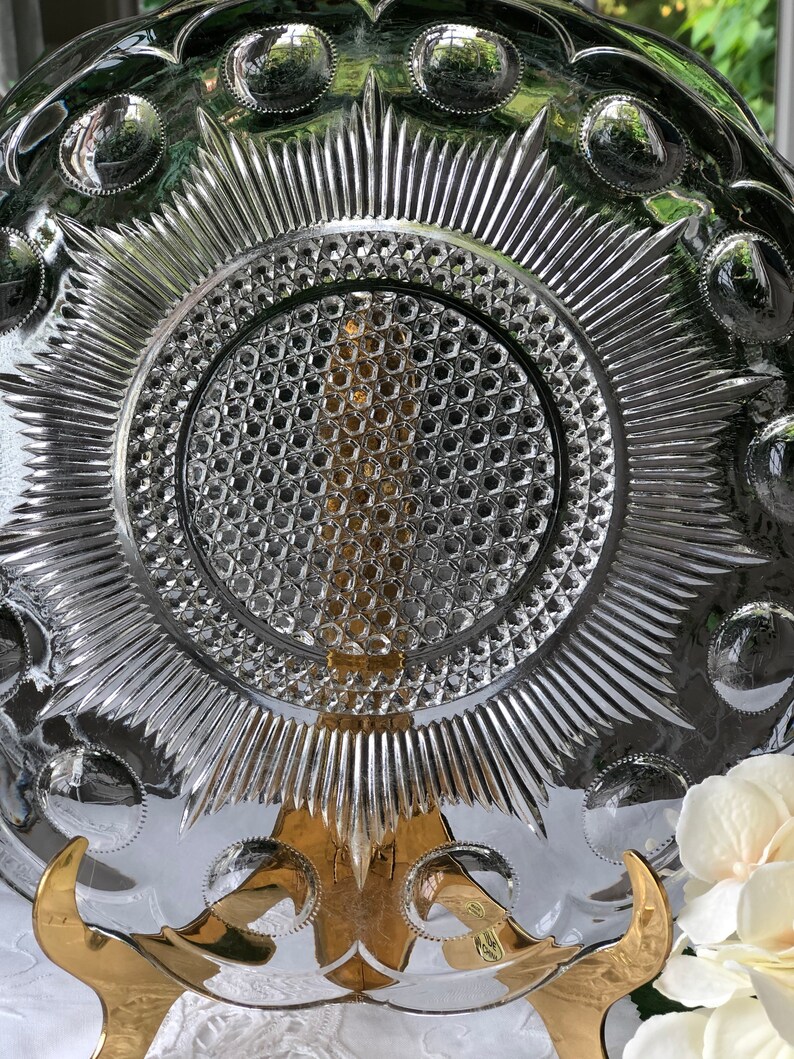 May include: A clear glass plate with a raised, starburst design and a central, circular, honeycomb pattern. The plate has a scalloped edge with raised circles around the rim.