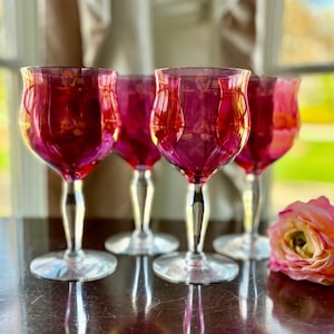 May include: Set of five wine glasses with a deep pink hue and clear stems. The glasses have a fluted design and are arranged on a dark wooden surface.