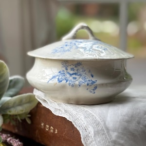 May include: Cream-colored ceramic serving dish with a lid, adorned with blue floral patterns. The dish has a curved shape and a handle on the lid. It rests on a vintage book and lace cloth, suggesting a vintage or antique aesthetic.