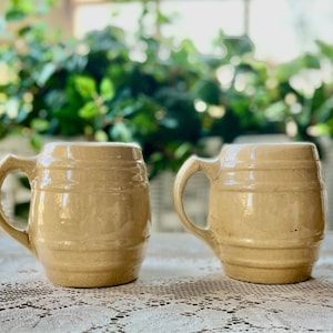 May include: Two vintage ceramic mugs with a light brown glaze. The mugs have a barrel shape and a handle. They are sitting on a white lace tablecloth.