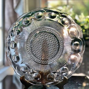 May include: A clear glass plate with a raised, circular design. The plate has a scalloped edge and is on a gold metal stand.