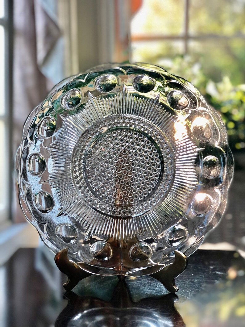 May include: A clear glass plate with a raised, circular design. The plate has a scalloped edge and is on a gold metal stand.