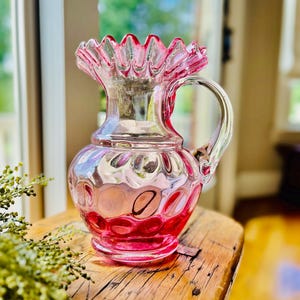 May include: A pink glass pitcher with a ruffled rim and a handle. The pitcher has a textured, bubble-like pattern on its body. The glass is a gradient of pink, with a darker shade at the base.