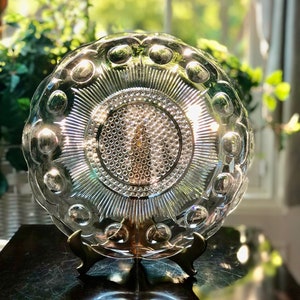 May include: A clear glass plate with a raised, textured design. The plate has a scalloped edge and a central design of concentric circles. The plate is on a gold metal stand.