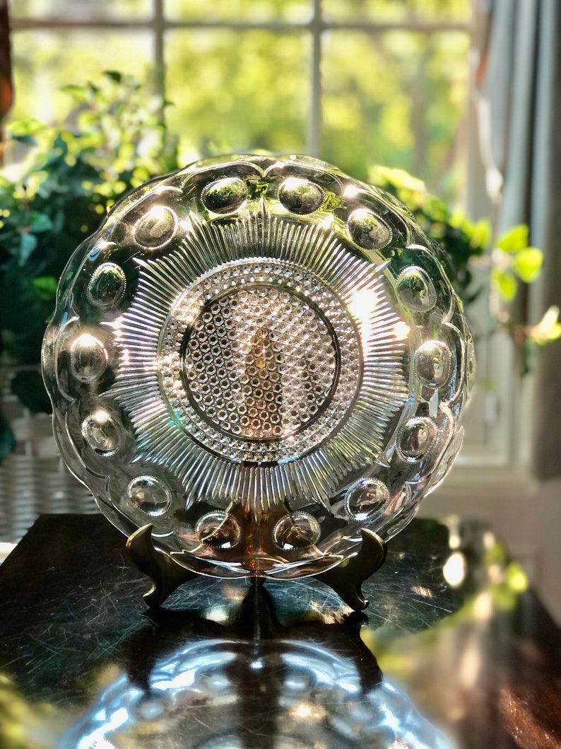 May include: A clear glass plate with a raised, textured design. The plate has a scalloped edge and a central design of concentric circles. The plate is on a gold metal stand.