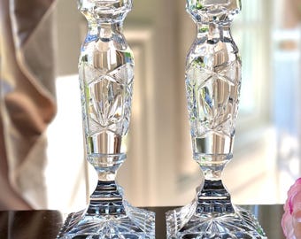 May include: Two clear crystal candle holders with intricate geometric patterns. Each holder has a square base, a detailed stem, and a top designed to hold a candle. The background is blurred, suggesting a room setting.