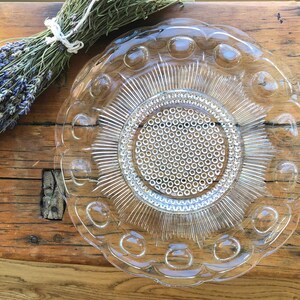 May include: A clear glass plate with a raised, textured design. The plate has a scalloped edge and a central circle with a repeating pattern of small circles.