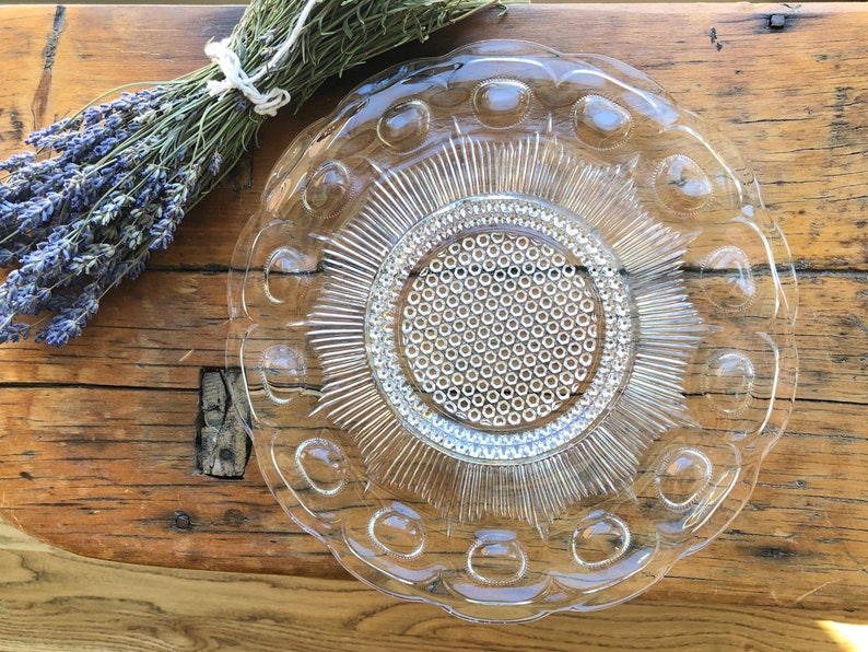 May include: A clear glass plate with a raised, textured design. The plate has a scalloped edge and a central circle with a repeating pattern of small circles.