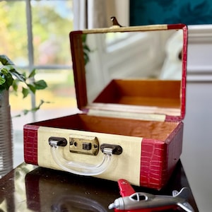 May include: A vintage-style vanity case featuring a cream-colored center panel and deep red crocodile-textured sides. The case includes a clear plastic handle, a brass-toned metal latch, and an interior mirror. A small red and silver toy airplane sits nearby.
