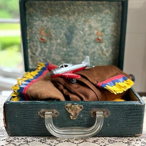 May include: An open vintage green suitcase with a brown blanket and a toy airplane inside. The blanket has yellow, blue, and red trim. The suitcase has a clear plastic handle and metal clasps. The interior of the suitcase has a patterned design.