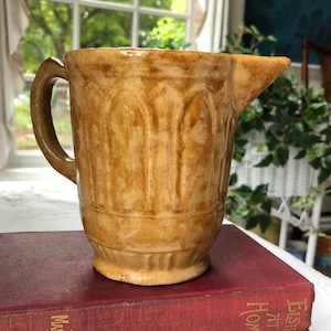May include: A brown glazed ceramic pitcher with a textured design. The pitcher has a handle and a spout.