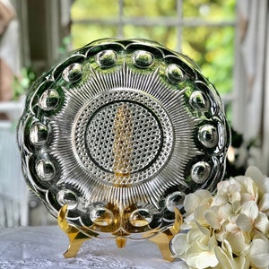 May include: A clear glass serving dish with a raised, textured design. The dish has a scalloped edge and a central, starburst pattern. The dish is displayed on a gold metal stand.