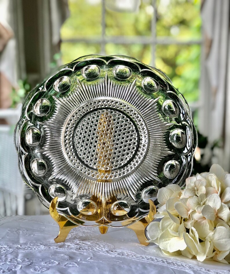 May include: A clear glass serving dish with a raised, textured design. The dish has a scalloped edge and a central, starburst pattern. The dish is displayed on a gold metal stand.