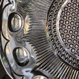 May include: A vintage glass serving dish with a raised, geometric pattern and a reflective surface. The dish has a scalloped edge and a series of raised circles around the center.