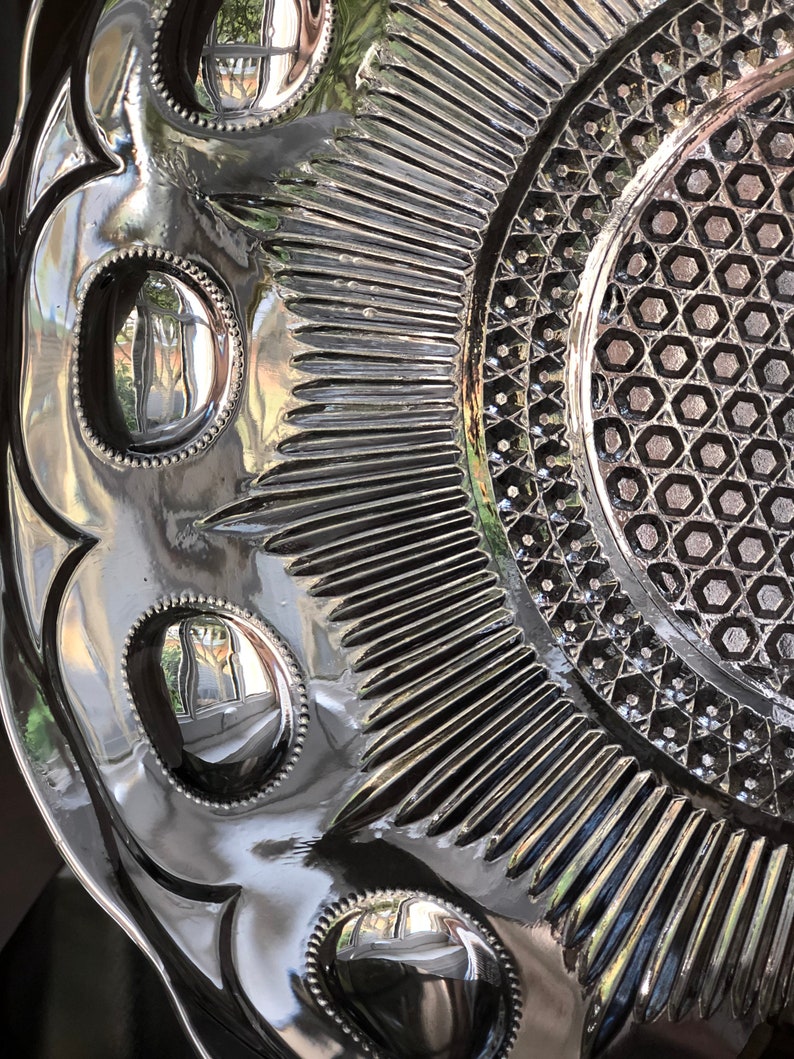 May include: A vintage glass serving dish with a raised, geometric pattern and a reflective surface. The dish has a scalloped edge and a series of raised circles around the center.