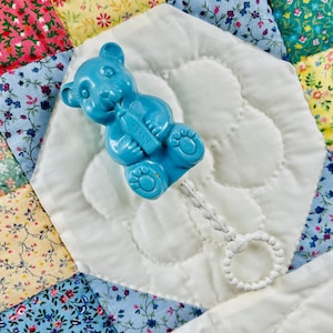 May include: A blue plastic baby rattle shaped like a teddy bear holding a bottle. The rattle has a white braided cord with a white plastic ring at the end. The rattle is on a white quilted blanket with a patchwork design.