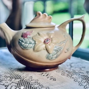 May include: A vintage ceramic teapot with a floral design. The teapot is decorated with a large yellow flower, green leaves, and pink accents. The teapot has a lid with a decorative knob.