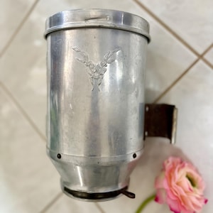 May include: A vintage, cylindrical metal container with a lid and a decorative emblem. The container is silver-toned with a dark brown bracket attached to the side. A pink flower with a green stem rests nearby.