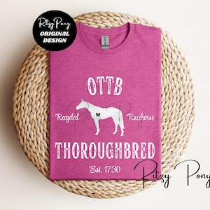 May include: A magenta t-shirt with white text and a horse graphic. The text reads "OTTB THOROUGHBRED Est. 1730" with "Recycled Racehorse" and "Ritzy Pony Original Design" branding. The shirt is displayed on a woven surface.