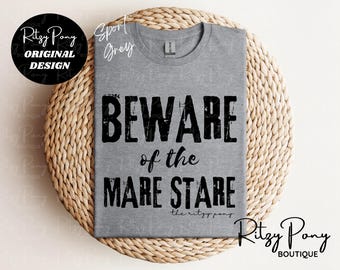 Può includere: T-shirt grigio sportivo con la scritta "BEWARE of the MARE STARE" in nero invecchiato. La maglietta presenta anche il testo "the ritzy pony" e il logo Ritzy Pony Boutique. La maglietta è esposta su una superficie tessuta.