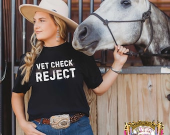 May include: A black t-shirt with the white distressed text VET CHECK REJECT, a beige cowboy hat, blue denim jeans, and a brown leather belt with a large ornate silver buckle. A grey horse stands in the background behind a wooden stall gate.