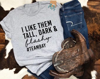 Puede incluir: Una camiseta gris con el texto "I LIKE THEM TALL, DARK & flashy #TEAMBAY" se muestra junto a unos vaqueros azules y botas de vaquero marrones. Las botas tienen costuras decorativas. Los artículos están colocados sobre una alfombra de piel de vaca.