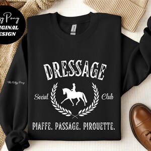 Pu&ograve; includere: Felpa girocollo nera con grafica bianca che recita "DRESSAGE Social Club" con un'illustrazione di cavallo e cavaliere all'interno di una corona d'alloro. Il testo "PIAFFE. PASSAGE. PIROUETTE." &egrave; sottostante. Design originale di Ritzy Pony.