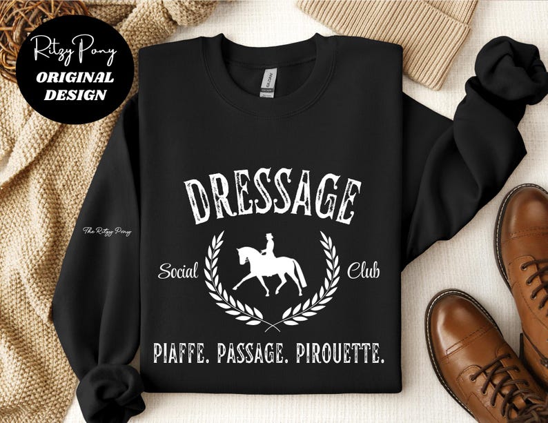 Pu&ograve; includere: Felpa girocollo nera con grafica bianca che recita "DRESSAGE Social Club" con un'illustrazione di cavallo e cavaliere all'interno di una corona d'alloro. Il testo "PIAFFE. PASSAGE. PIROUETTE." &egrave; sottostante. Design originale di Ritzy Pony.